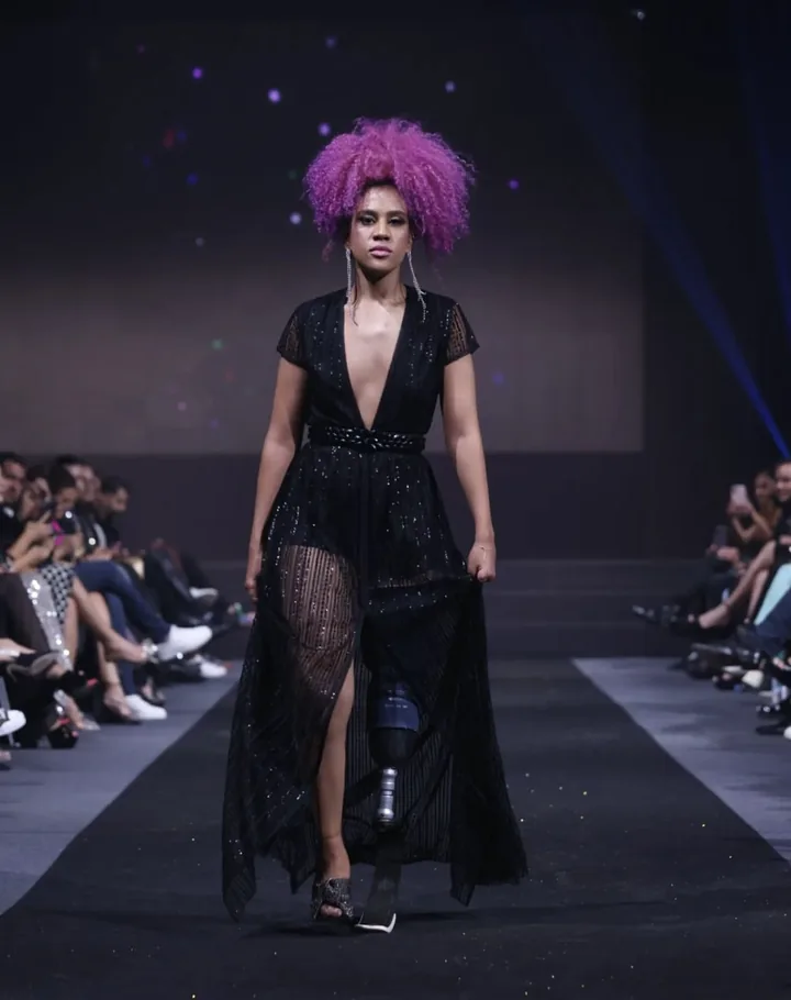 Zineb défile sur le podium, cheveux roses, robe noire, sa prothèse visible.